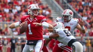 Two college football quarterbacks in action during live game competition, throwing football downfield with defensive pressure, intense athletic movement, stadium background with crowd, dynamic action shot