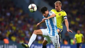 Professional football players in intense midfield battle, one wearing Argentina light blue and white striped jersey, other in Colombia yellow jersey, both competing for possession during international match, stadium background blurred, dynamic action shot showing athletic performance and competition intensity