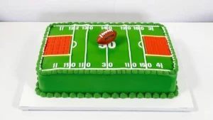 Professional rectangular football field cake with vibrant green frosting, white yard lines, 50-yard marker, end zone markings in team colors, small fondant football on the field, decorated on white cake board under soft studio lighting
