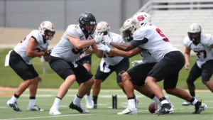 College football players executing high-intensity defensive drill, multiple athletes in action stance demonstrating proper tackling technique and alignment, outdoor practice field setting, athletic concentration visible