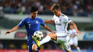 Professional male soccer players competing intensely during international match, one player in white jersey challenging another in blue jersey for possession, dynamic movement captured mid-play, stadium background blurred