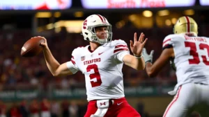 College football quarterback mid-throw during game, wearing Stanford Cardinal uniform, stadium lights, defensive pressure visible, athletic form and concentration