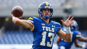 Athletic male college football player in Tulsa Golden Hurricane uniform executing perfect passing form during intense practice session, sweat visible, focused expression, professional stadium background