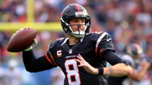 Professional football quarterback in throwing motion during game, wearing helmet and uniform, field blurred in background, dynamic athletic movement captured mid-throw